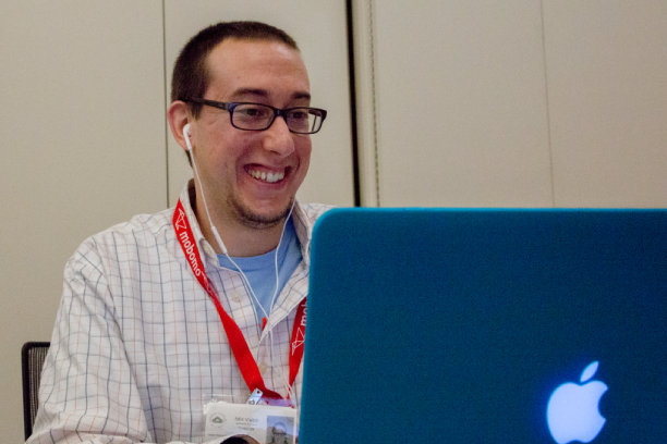 Man with glasses and a red lanyard smiles while using a blue Apple laptop.