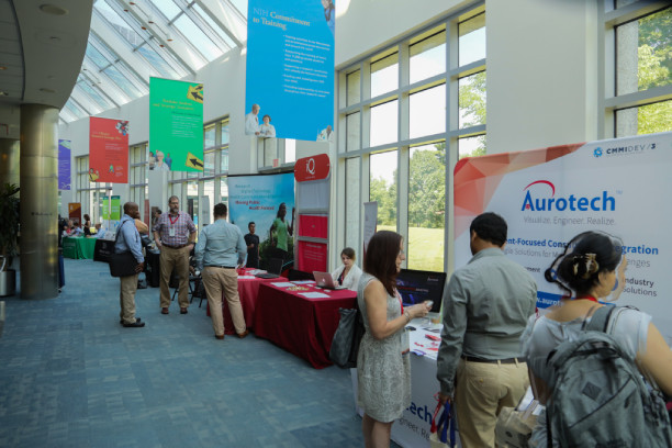 People interact at various booths in a well-lit convention center hallway with large windows.